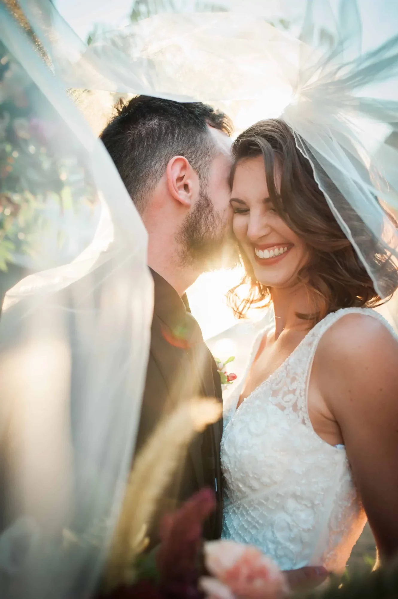 Groom whispers in bride's ear and making her laugh all while under the veil and the sun shinning through. Couple posing for a Bakersfield wedding photographer.